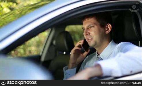Busy young businessman having a conversation with a business colleague using a smartphone while sitting in the driver's seat in a modern vehicle. Side view. Close-up. Professional businessman negotiating with a business partner via mobile phone while driving a car.