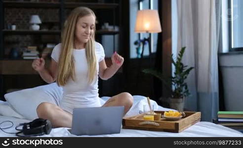 Carefree beautiful woman in white underwear sitting crossed legs on white bed sheet, listening to music with headphones during healthy morning breakfast in bed. Cheerful young female eating oatmeal, drinking orange juice and enjoying leisure at home.