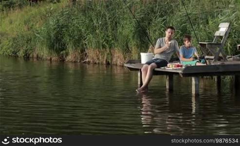 Carefree hipster father and teenage son sitting on a wooden pier, enjoying a meal while fishing together at a calm pond against a rural landscape background. Relaxed dad and boy having a snack and spending the weekend fishing on the lake. Slow motion.