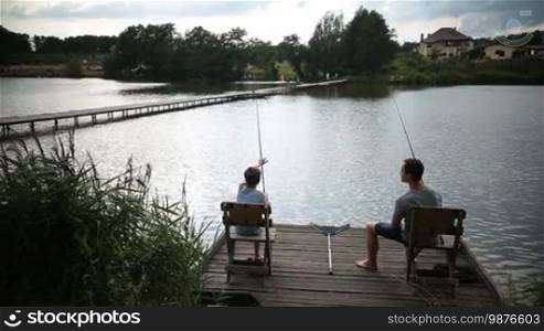 Carefree son and dad bonding with each other and talking while sitting on wooden pier chairs with fishing rods by the lake. Back view. Handsome father and teenage boy enjoying fishing at freshwater pond against rural landscape background.