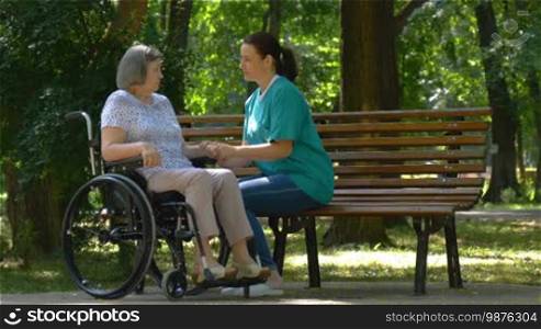 Caregiver talking to disabled senior woman in wheelchair outdoors in summer park.