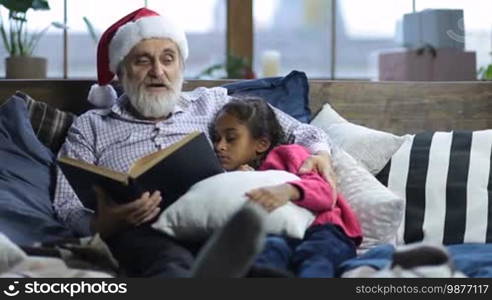 Caring grandfather with grey beard and in Santa hat reading a book to his sleepy cute mixed race granddaughter as they lie in bed on Christmas Eve. Affectionate grandpa cuddling his adorable girl while reading bedtime story at home on Christmas.