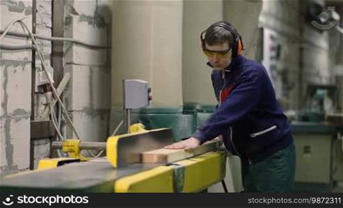 Carpenter working on electric buzz saw leveling boards. Skilled worker wearing safety glasses and earmuffs leveling wooden plank using milling machine in workshop.