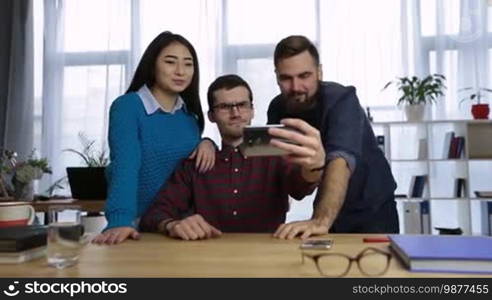Casual diverse business team taking self-portrait with smartphone in startup office. Smiling business people making funny facial expressions while taking selfie on mobile phone in office. Dolly.