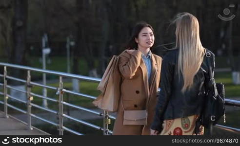 Charming Asian stylish woman holding shopping bags over her shoulder, standing on the pedestrian bridge while communicating with her girlfriend outdoors. Young female shopaholics chatting about latest gossips.