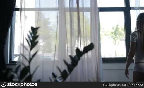 Charming blond woman with beautiful straight long hair in white underwear, t-shirt and panties, stretching near the window after early morning awakening. Back view. Stretching young sexy female exercising to get energy. Dolly shot.