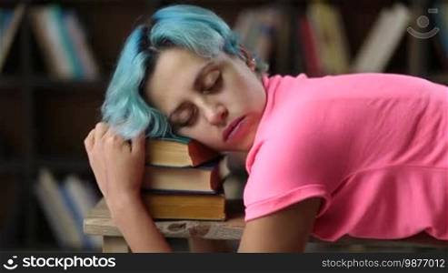 Charming hipster girl with blue hair sleeping on a pile of books in library. Side view. Exhausted of studying college female student napping on stack of books after reading tutorial material in university library.