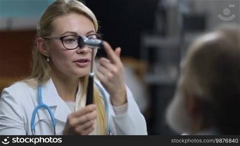Charming long blonde female neurologist in eyeglasses testing reflexes of senior male patient's eyes with neurological hammer during medical exam. Confident female doctor inspecting patient's nervous system using test hammer in clinic.