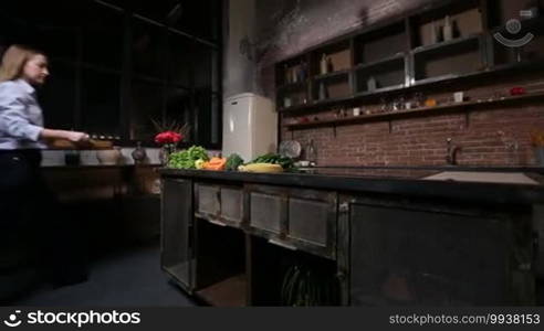 Charming woman preparing vegan smoothie with organic fruits and vegetables. Young lady bringing wooden tray with ingredients to the rustic styled kitchen and laying them out on the table to prepare a healthy vegetarian smoothie at home.