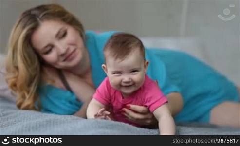 Charming young mother lying on her side on bed and encouraging her baby girl to crawl. Smiling infant child trying to crawl on bed with mother's help in bedroom. Mommy playing with little daughter while spending leisure together at home.