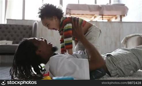 Cheerful African American father lying on the floor, holding his adorable toddler son and having fun. Smiling dad and his little child relaxing at home and fooling around while spending leisure together. Side view