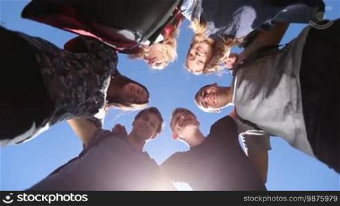 Cheerful college students standing in huddle, looking down at camera and smiling over blue sunny sky background. Low angle view. Positive teenage friends in circle looking down with radiant smiles.