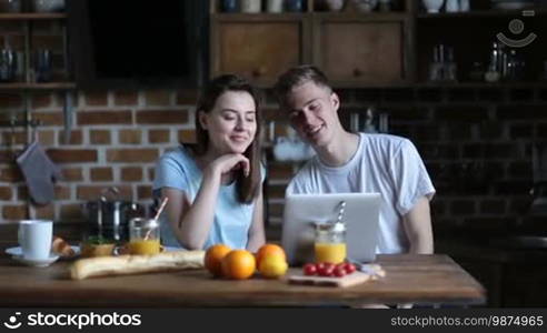 Cheerful couple video chatting via Skype using a laptop computer while sitting at the kitchen table and having breakfast in the morning. Positive young family communicating over a video call with parents via the internet at home.