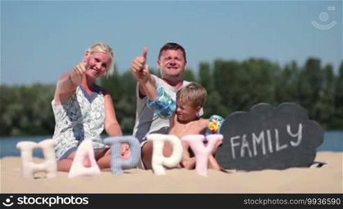 Cheerful family with son sitting on the beach and giving thumbs up during sea holidays. Foreground word happy made of fabric padded letters and word family written with chalk on black board