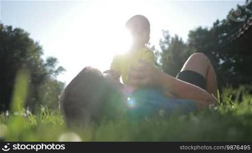 Cheerful father and his little infant baby boy playing outdoors in the glow of a beautiful sunset while spending leisure together in a summer park. Low view. Affectionate dad lying on his back with a smiling toddler son on his chest, having fun and relaxing on the lawn.