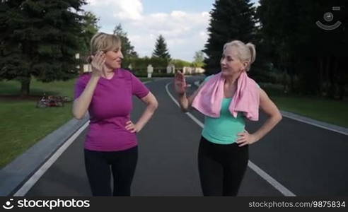 Cheerful fitness sport blonde women giving each other high five after running in park. Beautiful fit senior female friends celebrating a good jogging workout outdoors. Slow motion. Steadicam stabilized shot.