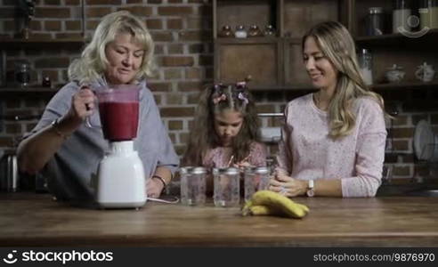Cheerful grandmother pouring fresh blended berry smoothie into mason jars while family sharing healthy breakfast together in the kitchen. Multi-generation family enjoying tasty healthy smoothie at home. Dolly shot. Slow motion.