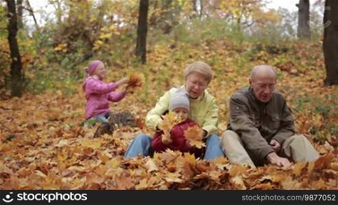 Cheerful grandparents and cute toddler grandson enjoying autumnal nature in park. Happy teenage girl in warm clothes collecting yellow maple leaves on background. Portrait of happy multi-generation family relaxing in autumn park.