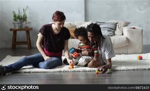 Cheerful multiethnic family with adorable little mixed race toddler son relaxing and playing with toy cars at home. Smiling pregnant Caucasian mother, handsome African American father with dreadlocks and their child sitting on the floor and playing with toys.