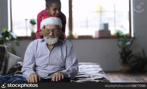 Cheerful small mixed race girl holding eyes of her grandfather closed as her younger sister giving Christmas gift box to grandfather. Joyful grandpa in Santa hat with white beard receiving Xmas present from adorable granddaughters on winter holidays.