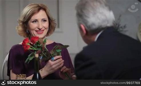Cheerful wife receiving bunch of red roses from her devoted husband on anniversary. Beautiful senior lady smelling the bouquet of flowers that she has just received on Valentine's day from her elderly man during romantic dinner at the restaurant.