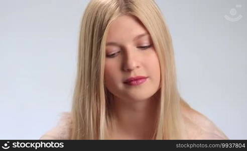 Cheerful young blonde woman slowly lifting up her head, looking mysteriously at the camera and smiling on white background. Positive teenage girl with radiant smile gazing at the cam closeup.