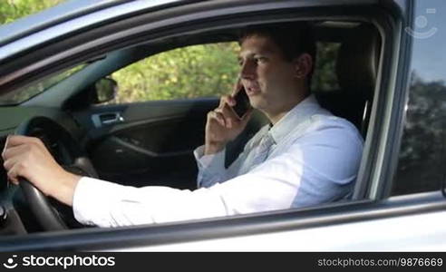 Cheerful young business executive in white shirt and tie talking on mobile phone as he sits in his car and holding steering wheel with one hand. Successful businessman communicating with business partner on smartphone while driving car. Slo mo. Stabilized shot.