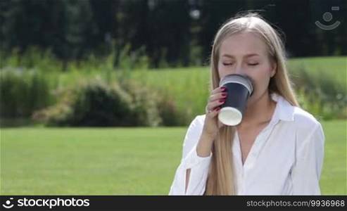 Cheerful young woman sitting on green grass outside in the park drinking coffee and resting in the nature during coffee break