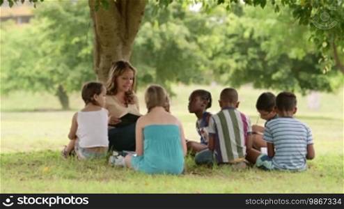 Children and education, young woman at work as educator reading book to boys and girls in park