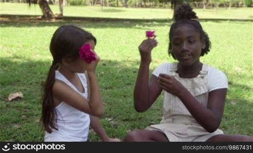 Children and fun, two female friends talking, laughing and playing with flowers outdoors in a city park