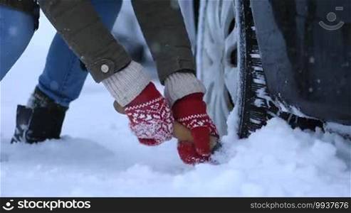 Close up female hands holding shovel and removing snow from front wheel during snowstorm in winter. Woman's hands in red woolen mittens shoveling and removing snow from her stuck car.