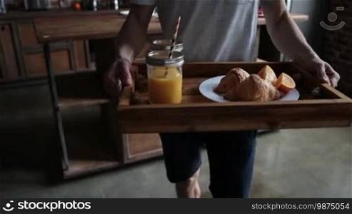 Close-up midsection of a young man carrying breakfast on a wooden tray. Male hands holding a wooden tray with orange juice in mason jars, slices of orange, and croissants to surprise his beloved woman with breakfast in bed. Slow motion. Steadicam stabilized shot.