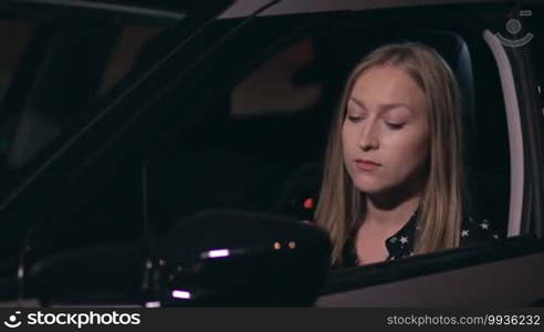 Close up of a pretty woman driver applying red lipstick and looking at the car's side rearview mirror while standing on a roadside of the city street at night.