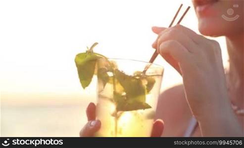 Close up of a woman enjoying a mojito cocktail at the seaside at sunset sipping it through a straw on her summer vacation