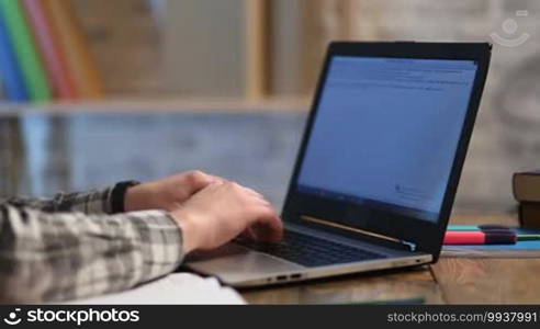 Close up of male student hands typing text on modern laptop on wooden table while studying at home. Young teenager working on computer in library and preparing for exams. Hipster using digital gadget.