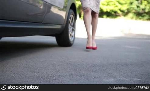 Close-up perfect woman's legs in red high heels coming to the parked car. Fashionable female driver getting in luxury car. Elegant lady with perfect long legs wearing high heel shoes stepping into automobile. Dolly.