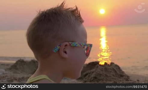 Close-up shot of a blond little boy in colorful sunglasses on a background of a quiet sea and golden sunset. Summer holidays