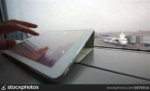 Close-up shot of a female hand typing on a tablet computer by the window at the airport with a boarding plane in the background