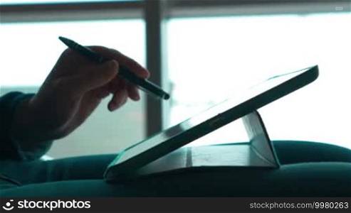 Close-up shot of a female hands working with a tablet computer using a pen to touch the screen. She is holding the tablet PC on her lap against the window with bright sunlight