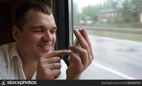 Close-up shot of a happy young man typing a text message while sitting by the window on the train