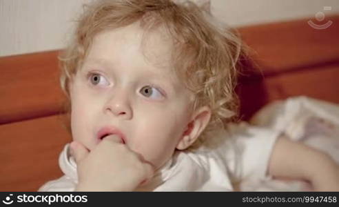 Close-up shot of a little boy on a bed looking somewhere with his fingers in his mouth