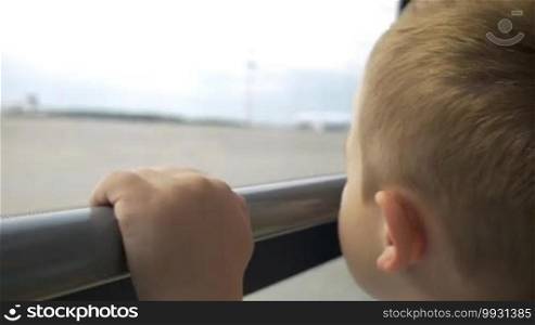 Close-up shot of a little curious boy looking out the window of a bus moving on the take-off strip at the airport. Time before departure