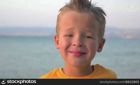 Close-up shot of a little cute boy with mohawk hairdo smiling. Portrait against blue sea and sky background