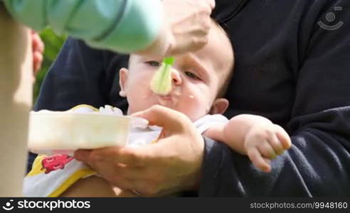 Close-up shot of a mother feeding a baby with cereal while the father is holding him. Breakfast outdoors