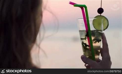 Close-up shot of a woman having a refreshing mojito cocktail with a straw and looking at the sea. Holidays and summer vacation