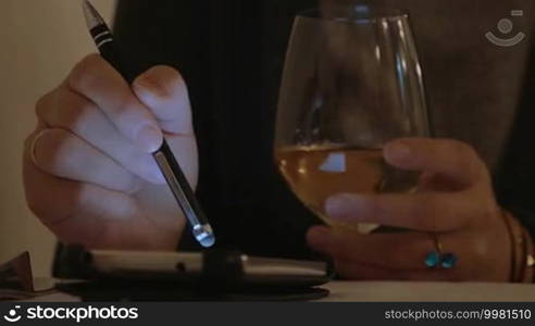 Close-up shot of a woman in a cafe drinking white wine and using a smartphone with a pen