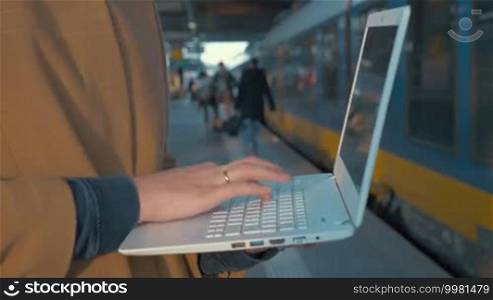 Close-up shot of a woman typing on a laptop at the station. She is standing on the platform by the train at a stop