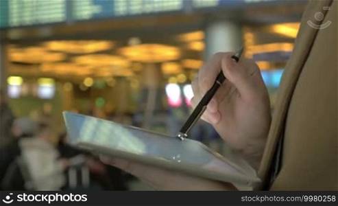 Close-up shot of a woman typing on a tablet PC with a pen at the airport. She is sending a message while standing by the flight schedule