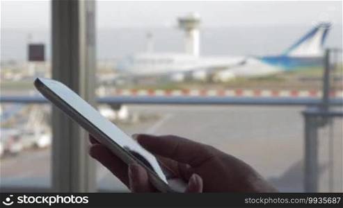 Close-up shot of a woman typing SMS on a smartphone by the window of an airport terminal with a view of a moving airplane