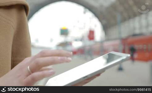 Close-up shot of a woman working with a tablet computer at the station. Defocused platform with a train and people in the background